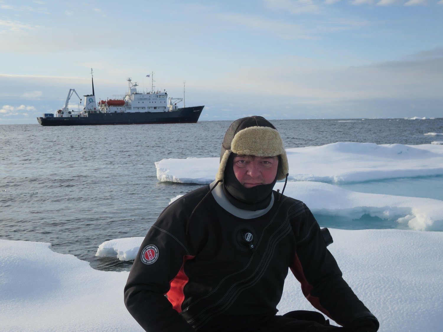 Diver in dry suit on Arctic ice with expedition vessel behind
